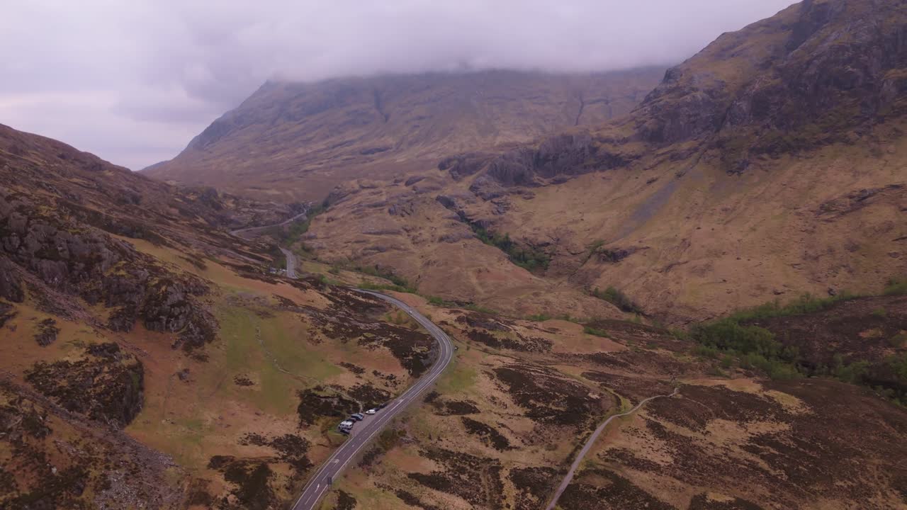Acercamiento aéreo de un coche conduciendo a través del espectacular valle de Glencoe en Escocia, rodeado de montañas, nubes y terreno accidentado