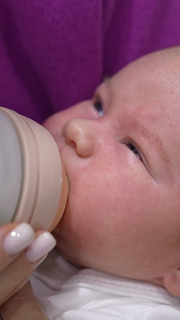 Newborn baby fed from a bottle lying in mother's hands. Baby boy screwing up his eyes against the light while being fed. Close up. Vertical video