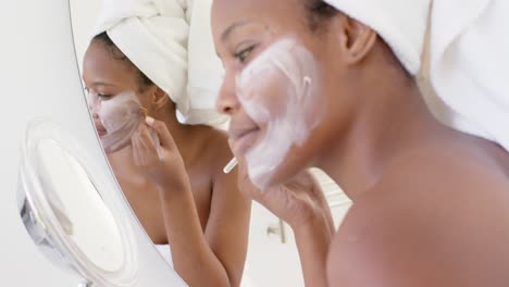 happy biracial woman with towel on head making mask on face in bathroom, slow motion