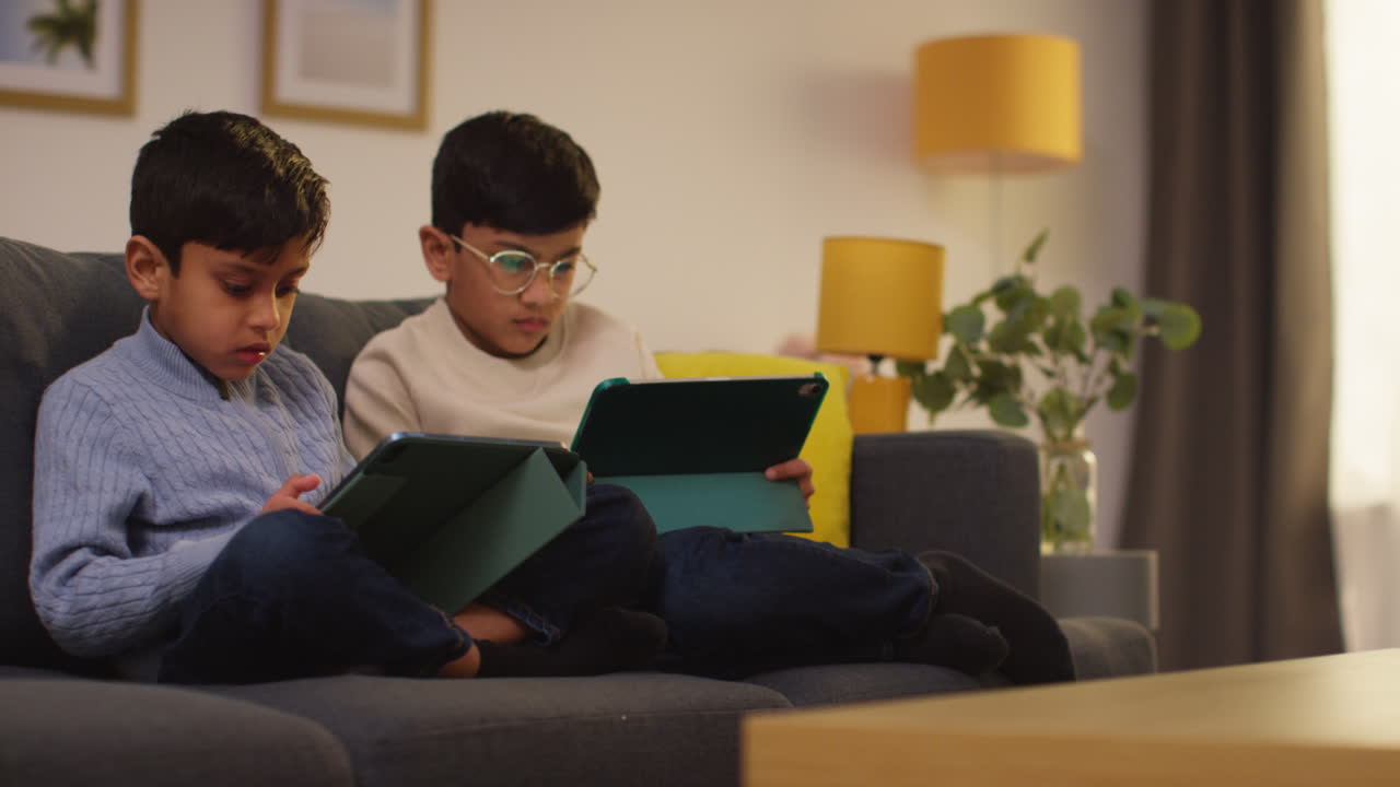 Two Young Boys Sitting On Sofa At Home Playing Games Or Streaming Onto Digital Tablets 14