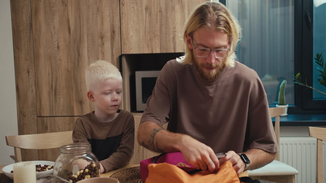 A blond man in glasses with a beard helps his little albino son with white hair pack things into a bright backpack in the kitchen after breakfast before the start of the school day