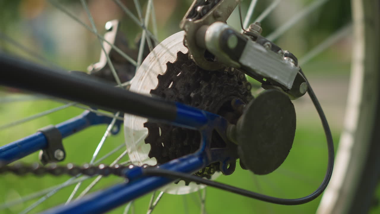 바가 천천히 회전하면서 자전거의 뒷바와 기어 시스템의 클로즈업, 체인과 바가 초점에 있으며, 배경에는 흐릿한 사람들이 고 초록색이 포함되어 있습니다.