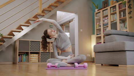 young fit woman sits cross legged on yoga mat at home stretching with arms out