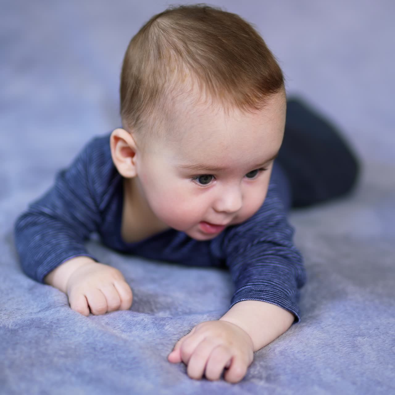 Nice Caucasian baby boy lying on his tummy on big bed. Mom's hand reaches the son and he responds with a cute smile. Grey backdrop in blur