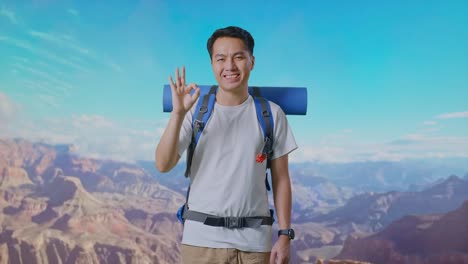 asian male hiker with mountaineering backpack smiling and showing okay gesture to camera while traveling at the top of mountain