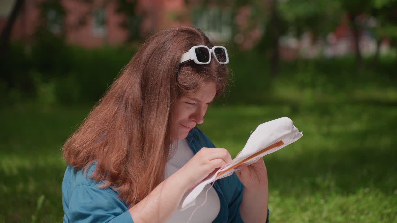 Woman deeply focused on embroidery work in bright sunlight, holding fabric tightly and stitching with precision, seated outdoors surrounded by green grass