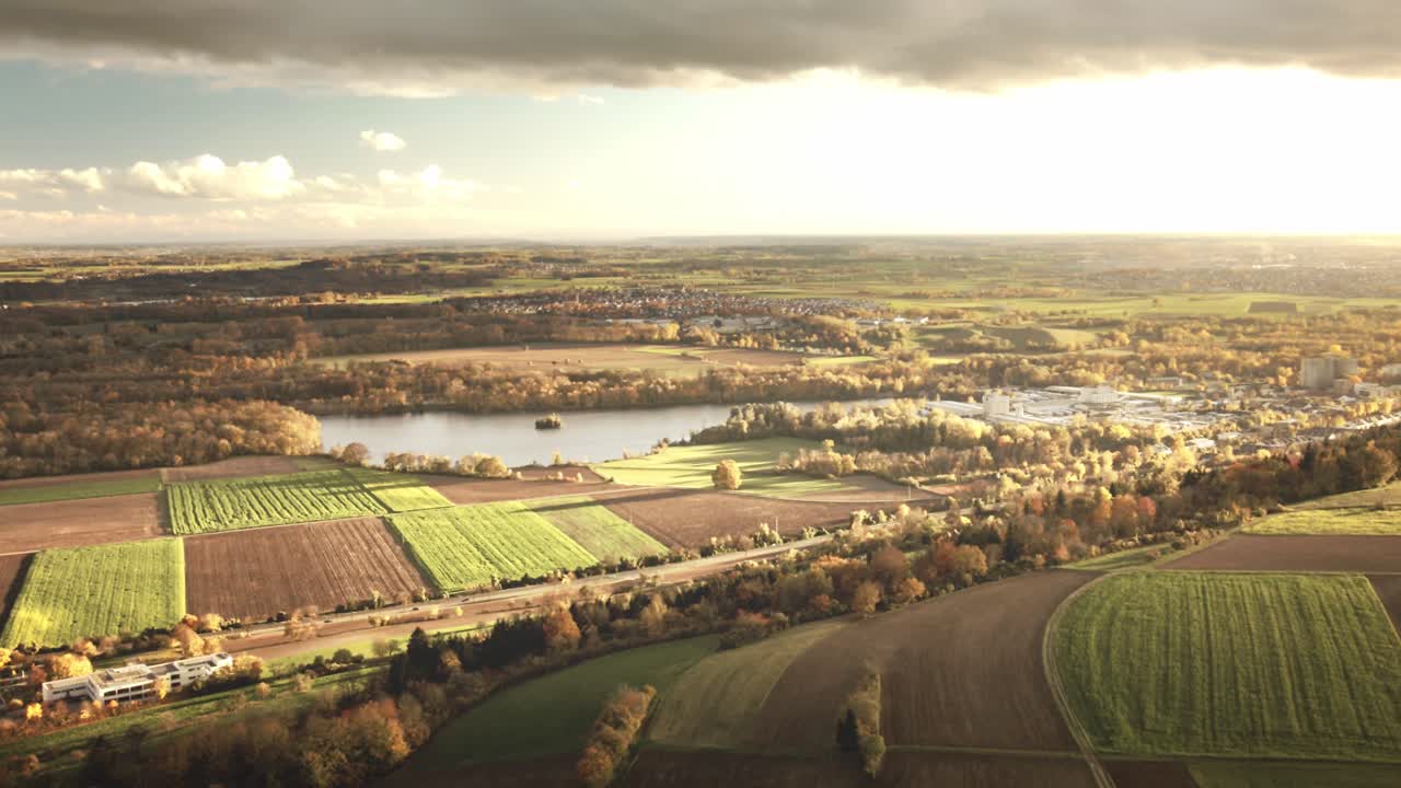 Camera glides forward above rural fields glowing in evening light, unveiling a winding lake, distant forests, and scattered villages under dramatic clouds
