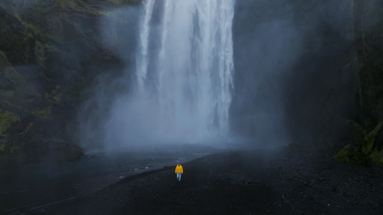 A Man In Yellow Jacket Is Walking Near Cascading Over Dramatic Black Rock Cliffs In Sk&oacute;gafoss, Iceland
