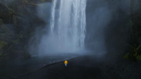 a man in yellow jacket is walking near cascading over dramatic black rock cliffs in skógafoss, iceland