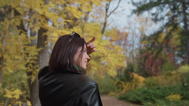Donna di colore con giacca di pelle che percorre un sentiero autunnale tra alberi dorati, si volta per toccarsi i capelli intrecciati, vista posteriore che trasmette uno stato d'animo rock contemplativo con colori cinematografici, quieta solitudine e stile audace