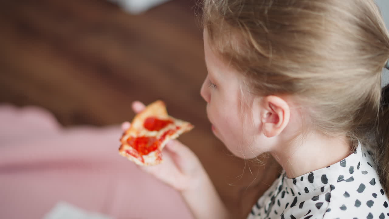 Young girl wearing spotted clothes with hair tied back bites into slice of pepperoni pizza while seated indoors, enjoying delicious snack in relaxed cozy environment