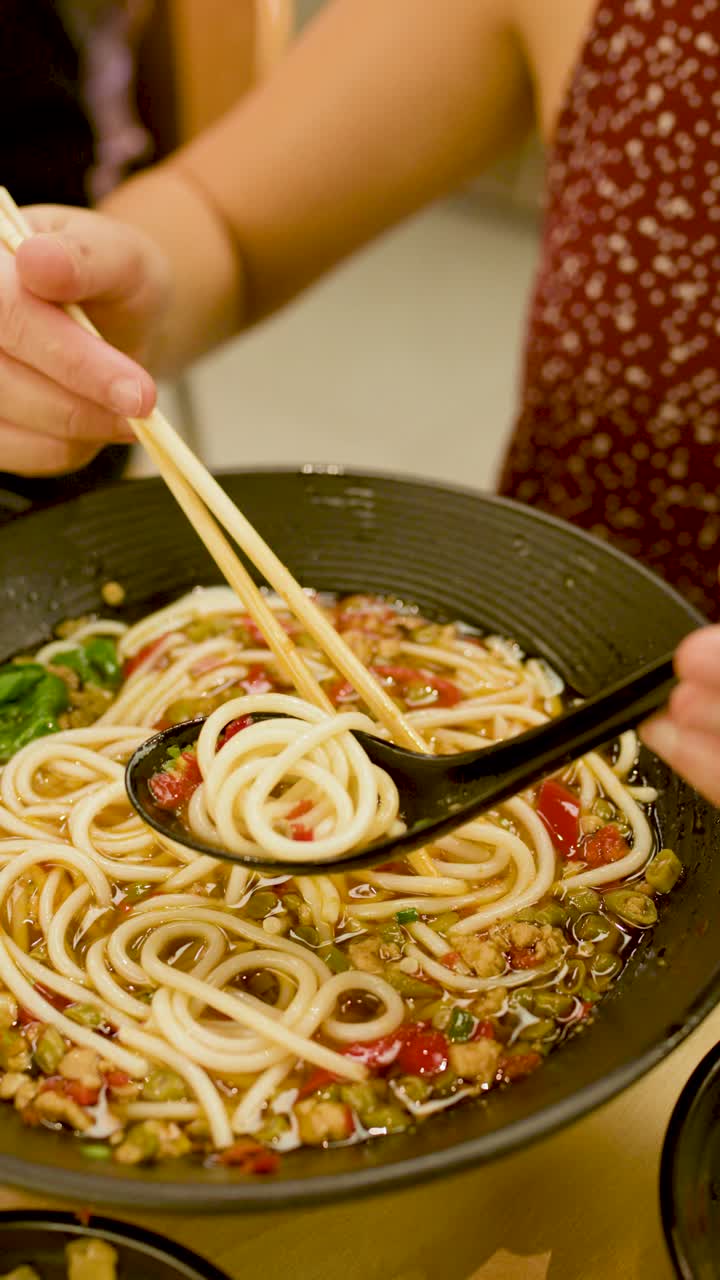 Una persona usa bacchette e un cucchiaio per sollevare una zuppa di noodle piccante in un ambiente da pranzo illuminato calorosamente.
