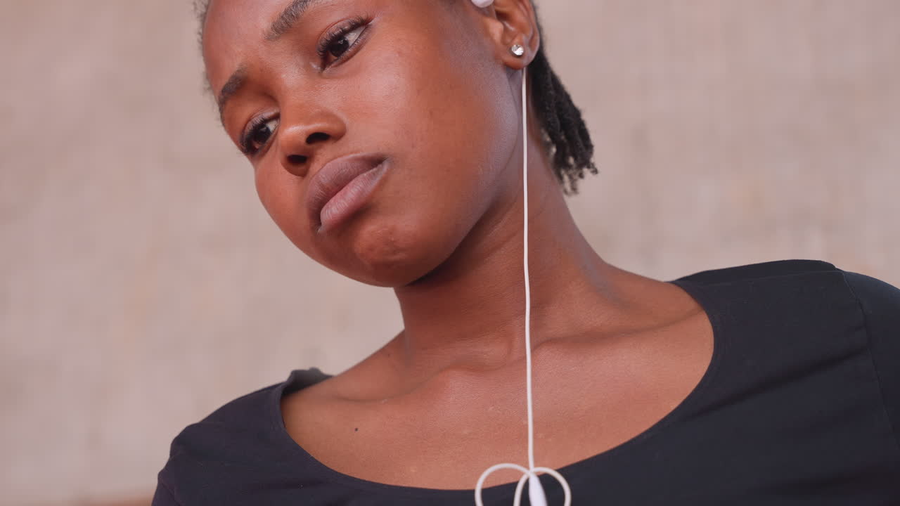 Donna di colore che ascolta gli auricolari, ritratto introspettivo che mette in risalto i capelli a trecce, un leggero sudore sulla pelle, sfondo neutro, una leggera inclinazione della testa e uno sguardo pensoso che suggeriscono una studentessa o una pendolare.