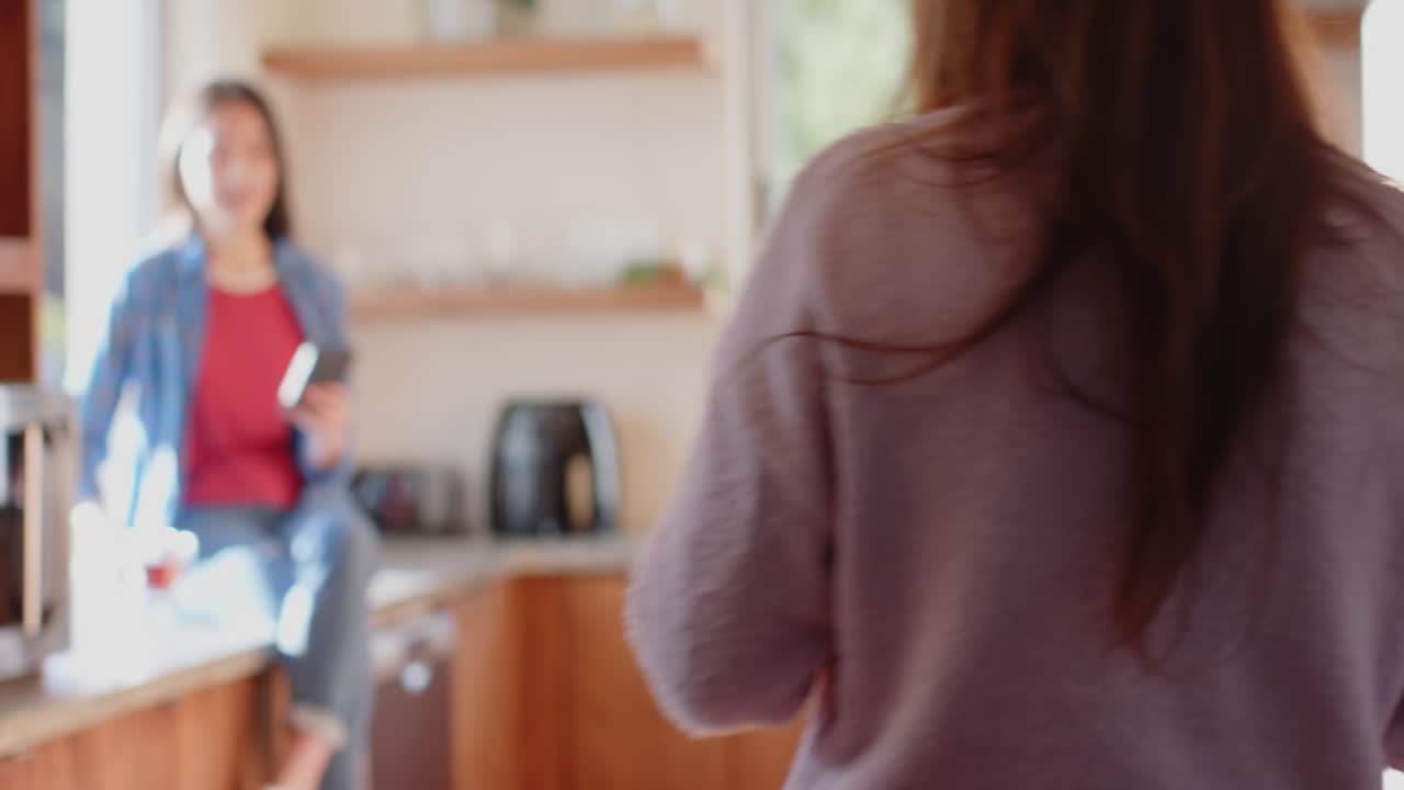 Talking and using smartphone, woman sitting on kitchen counter with another woman, copy space