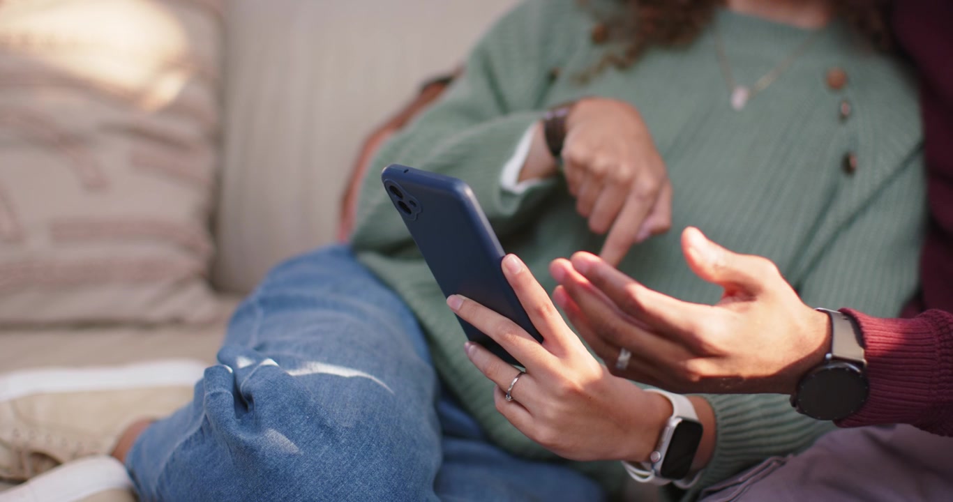 Pár sedící na gauči, žena ťukající do telefonu s modrým pouzdrem, muž natahující se, ukazující a sdílející obsah.