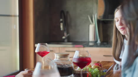 young woman in red shirt sits at wooden table with glass of red drink and food, surrounded by cozy kitchen atmosphere, enjoying quiet moment and pleasant time with friends in natural daylight