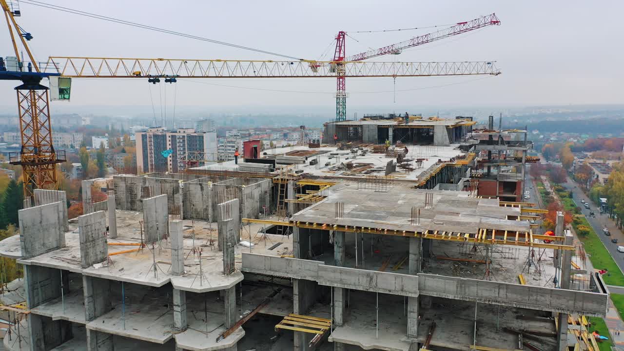 New construction site of shopping center. Aerial view on roof with workers and big cranes during construction on the city background. Drone moves back.