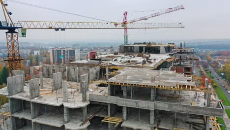 new construction site of shopping center. aerial view on roof with workers and big cranes during construction on the city background. drone moves back.