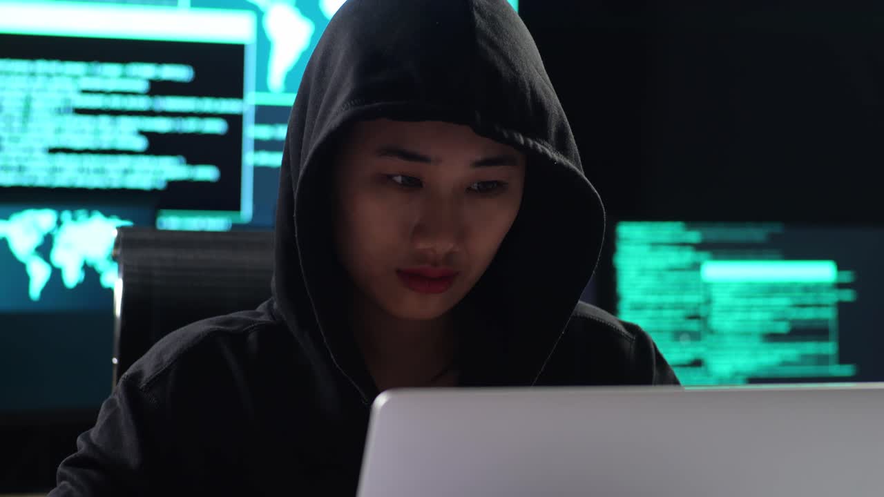 Close Up Of Asian Female Hacker In A Hood Drinking Coffee While Works On A Laptop With Maps And Data On Display Screens Of Multiple Monitors In A Dark Office Room