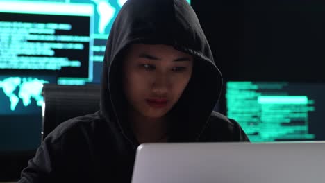 close up of asian female hacker in a hood drinking coffee while works on a laptop with maps and data on display screens of multiple monitors in a dark office room