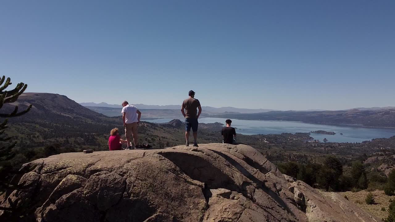 la gente está parada en un acantilado rocoso, rodeada de un paisaje natural intacto durante el día soleado, revelando un complejo aéreo a natural con lago y montañas