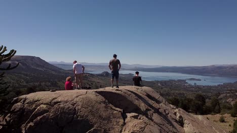 People-are-standing-on-a-rock-cliff,-surrounded-by-untouched-natural-landscape-during-sunshine-day,-revealing-aerial-to-natural-resort-with-lake-and-mountains