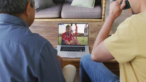 Video-Von-Vater-Und-Sohn,-Die-Auf-Der-Couch-Sitzen-Und-Sich-Ein-Fußballspiel-Auf-Dem-Laptop-Ansehen