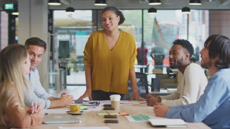 Standing-Businesswoman-Leading-Meeting-With-Colleagues-Sitting-Around-Table-In-Modern-Office