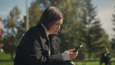 lady seated outdoors, focused on phone in her hand, with ring-adorned finger, and blurred background featuring greenery and trees on a bright sunny day