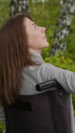 cheerful female sits in wheelchair in green park. young woman smiles opening palm to catch raindrops. positive person with injury backside view