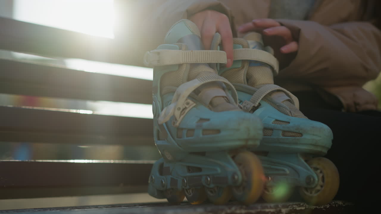 Close-up shot of a person walking to a park bench and sitting down, placing their rollerblades beside them. Sunlight filters through the scene, highlighting the details of the rollerblades