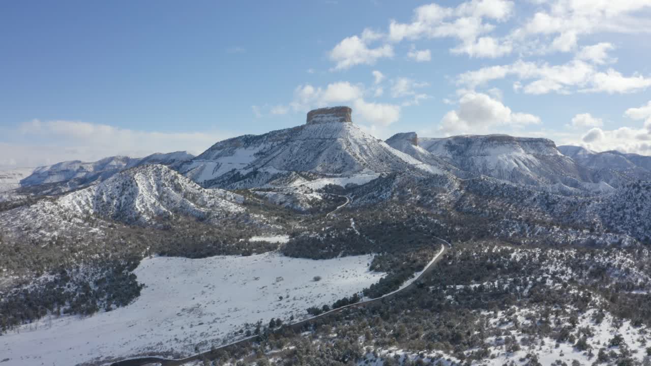 무인 항공기 영상은 화창한 날 드문 드문 녹지로 덮인 날카로운 봉우리가 있는 눈 덮힌 산맥을 여행하는 도로를 보여줍니다.