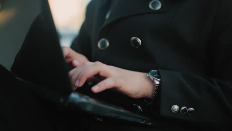 close up of individual hand in black suit operating laptop outdoors with attention to detail, featuring silver buttons, laptop ports, wristwatch, and soft bokeh background