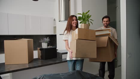 a happy brunette girl in a white t-shirt and jeans together with her happy brunette boyfriend with stubble walk into their new apartment with boxes in their hands and put them on a closed sofa in a new studio apartment modern