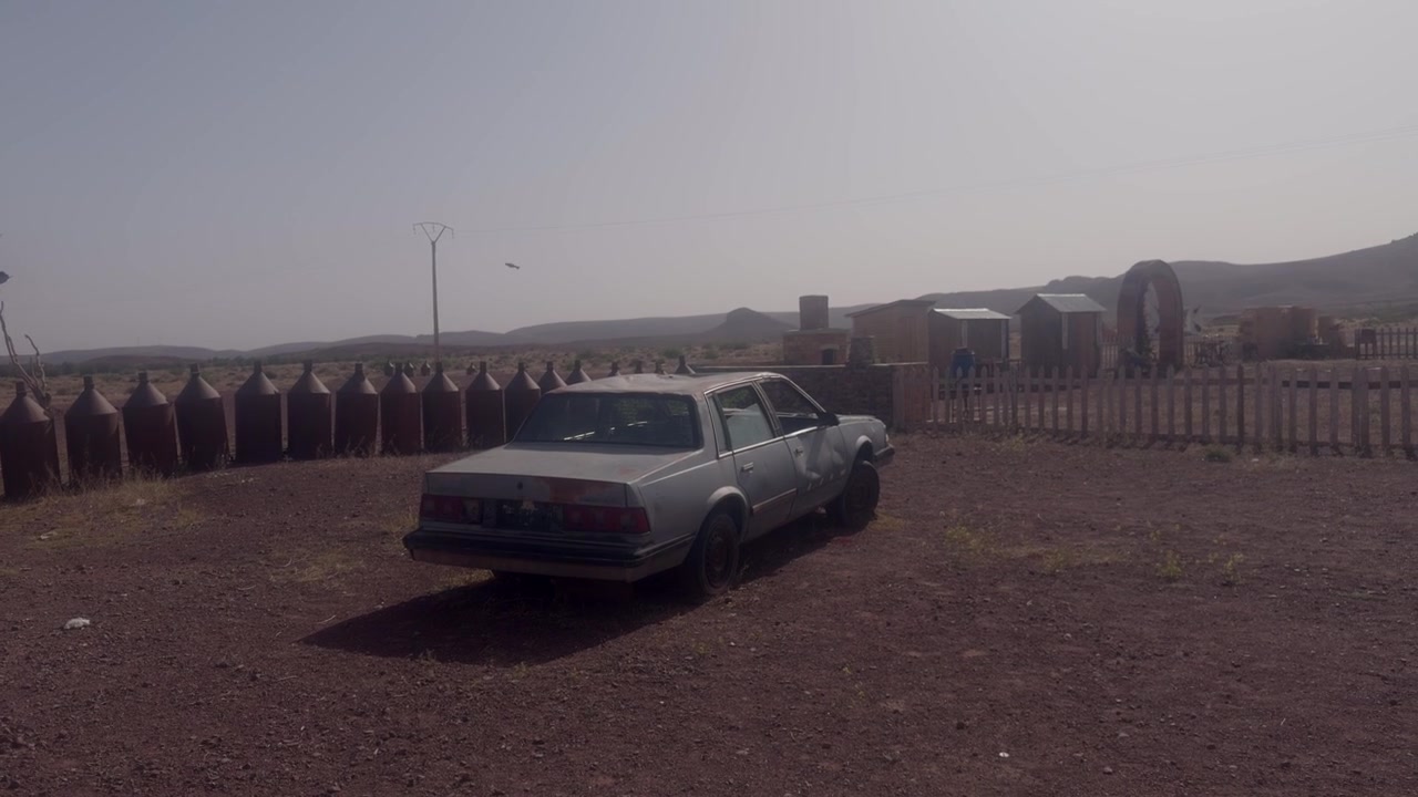 Sedán plateado viejo y desgastado estacionado en tierra junto a una valla oxidada en una gasolinera abandonada en el desierto, Marruecos.