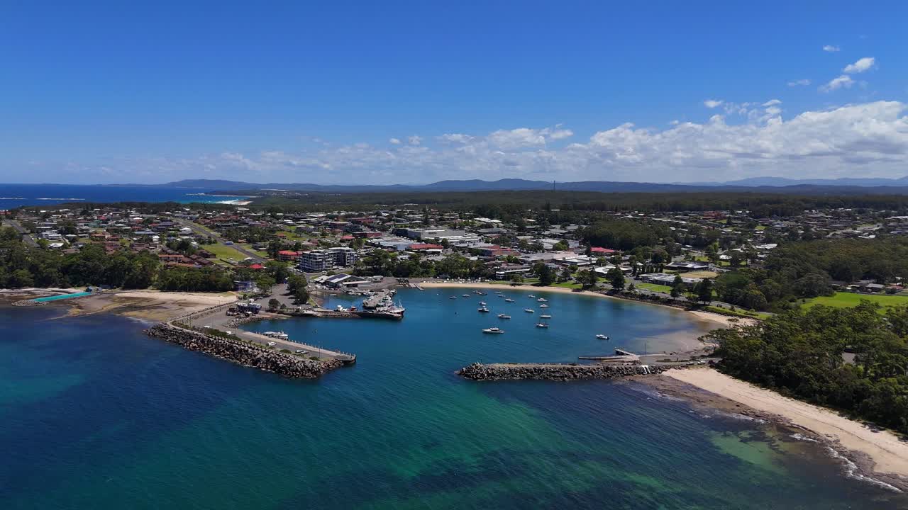 Aerial pullback establishing Ulladulla marina showing boats, docks, and sheltered harbour waters, bright blue sky