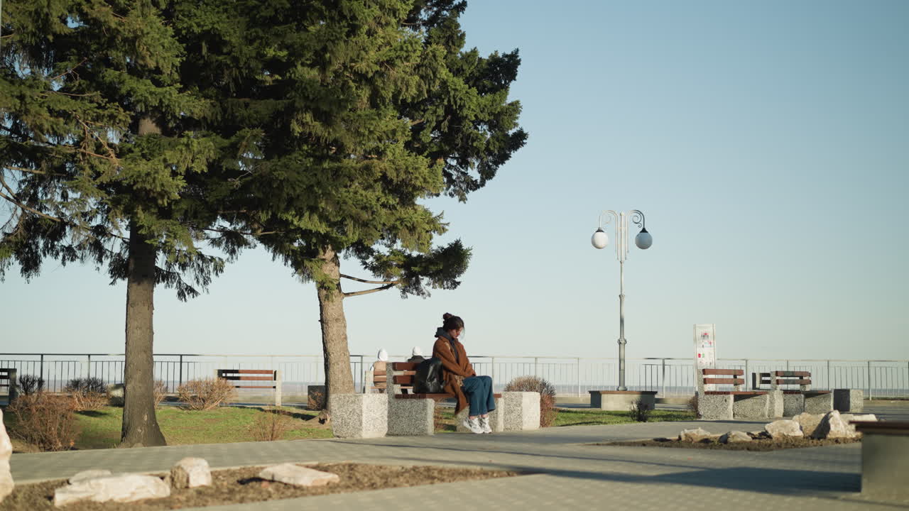 una mujer joven con un abrigo marrón, pantalones vaqueros azules y zapatos blancos se sienta sola en un banco en un parque urbano, su mochila negra a su lado. parece de mal humor y profundamente pensativa, con una expresión sombría