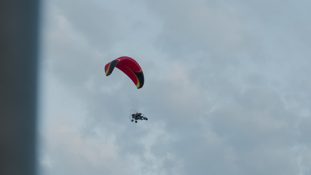 Silhouetted people flying powered paraglider trike under pastel evening sky, red canopy arched above while engine cage and wheels hover against soft cloud backdrop
