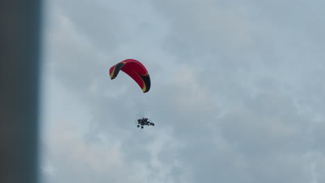 silhouetted people flying powered paraglider trike under pastel evening sky, red canopy arched above while engine cage and wheels hover against soft cloud backdrop