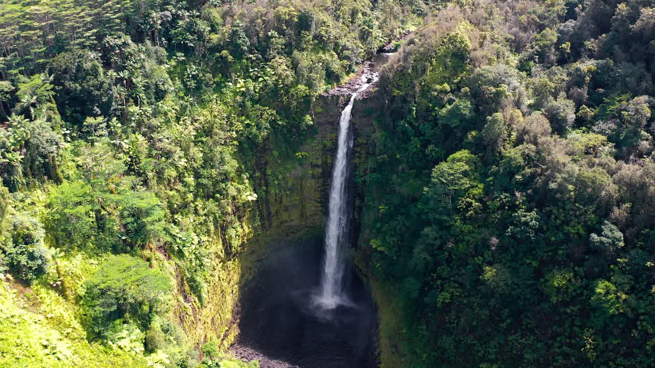A powerful waterfall streams down a towering rainforest cliff into a dark volcanic crater, its mist rising into the air, emphasizing the raw force of tropical nature
