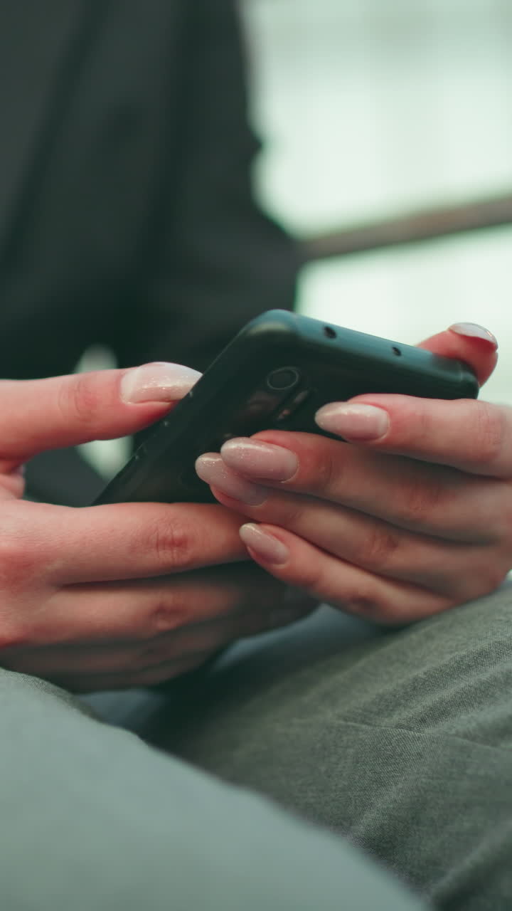 Close up of person with artificial nails using smartphone while seated outdoors, wearing black blazer and grey pants, with soft focus glass building in background