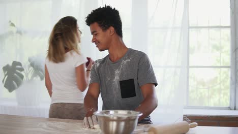 atractivo africano preparando masa en la mesa de madera. su novia se acerca, ofreciéndole la manzana pero tomándola