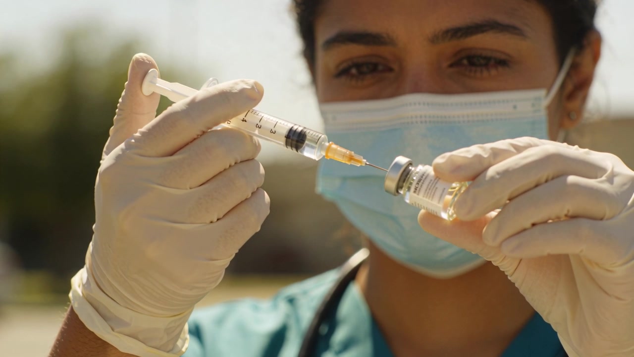 Holding vial and syringe, nurse drawing vaccine dose at outdoor site for vaccination in teal scrubs