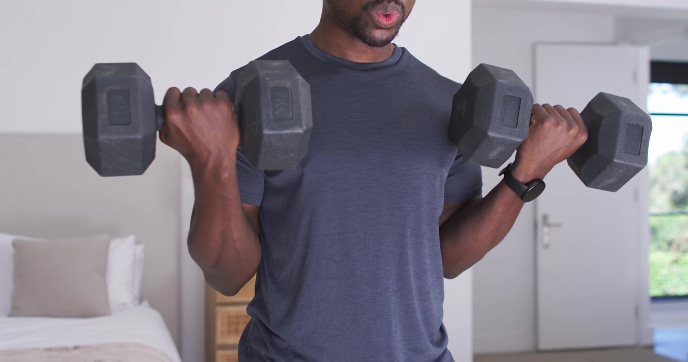 Adult African man starting bicep curls with hex dumbbells in bedroom wearing tee, building strength
