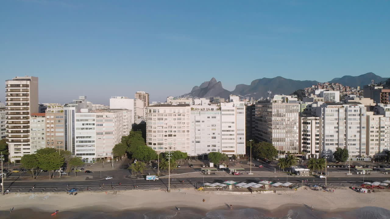 inclinación lenta aérea hacia abajo con la montaña de los dos hermanos e ipanema en el fondo, revelando las olas rodando en la playa de copacabana y la arquitectura del barrio en primer plano