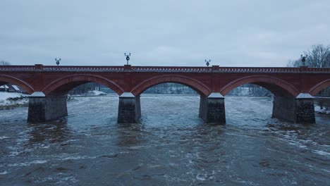 vista aérea de los rápidos del río venta durante la inundación de invierno, viejo puente de ladrillo rojo, kuldiga, letonia, día de invierno nublado, amplia toma ascendente de drones que avanzan