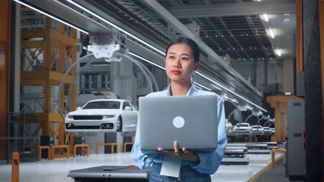 una trabajadora profesional asiática de pie con su portátil en una fábrica de automóviles ev, una instalación industrial de alta tecnología, observa mirando a su alrededor antes de concentrarse con su computadora portátil