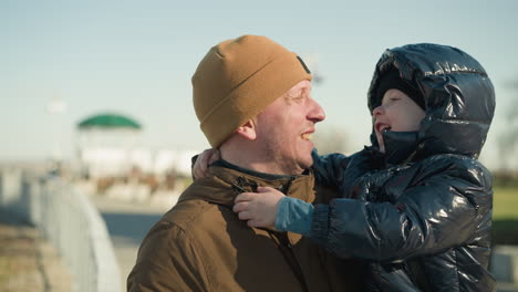 a father carries his son as the child playfully grabs his attention, creating a moment of joyful interaction, the father is wearing a brown jacket, and the son is dressed in a black puffy jacket