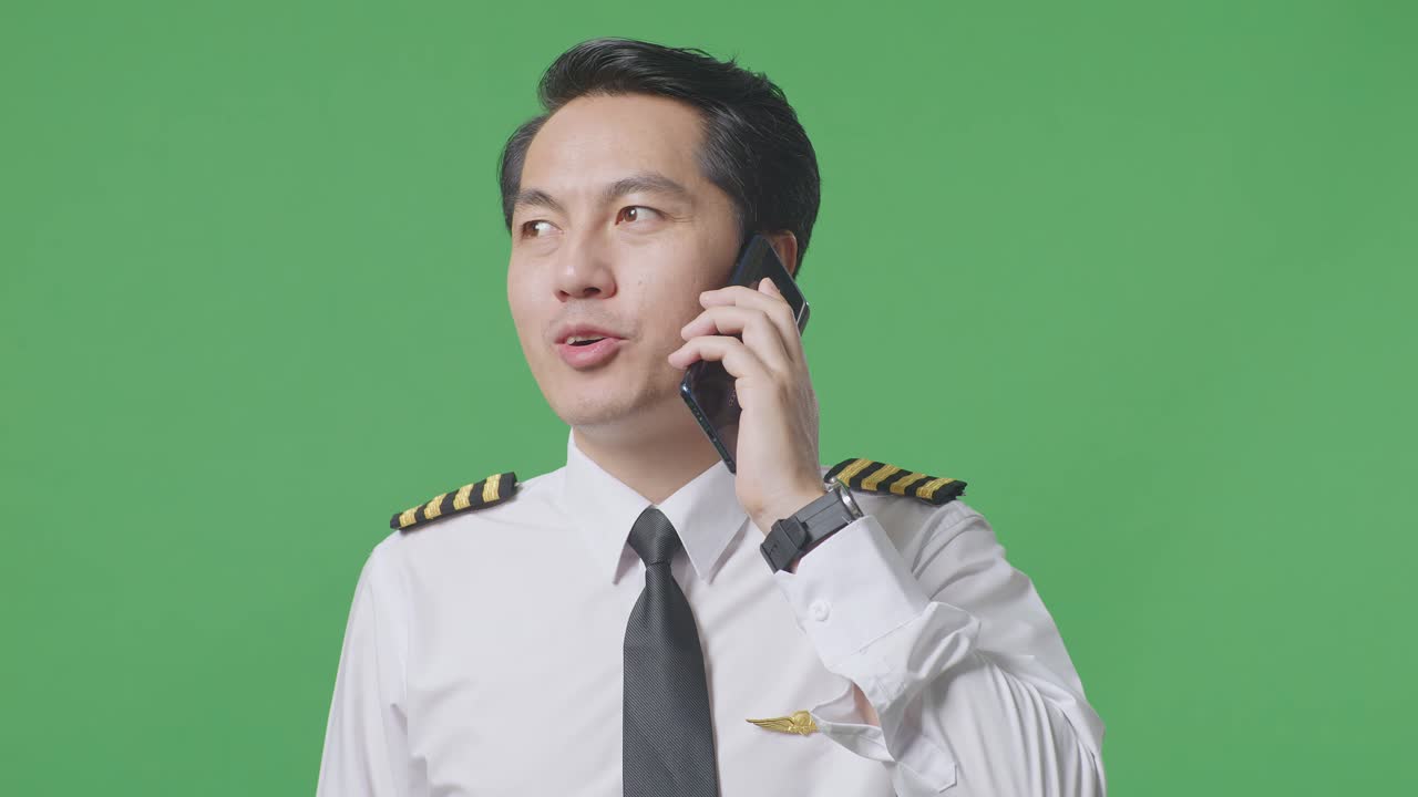 녹색 스크린 배경 스튜디오에 서서 스마트폰으로 대화하고 주변을 둘러보는 아시아 남성 조종사의 클로즈업