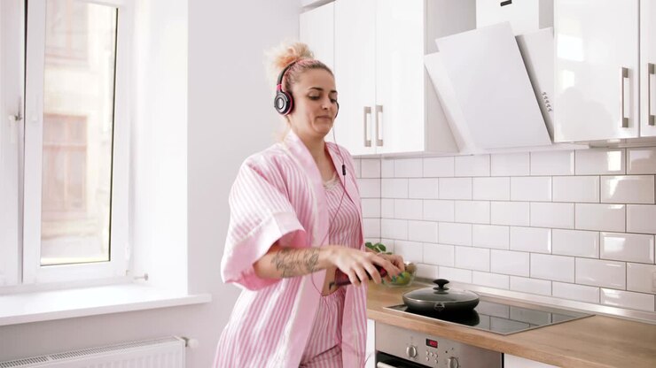 Giovane donna felice con i capelli rosa che balla in cucina, indossando vestiti rosa e ascoltando musica con le cuffie